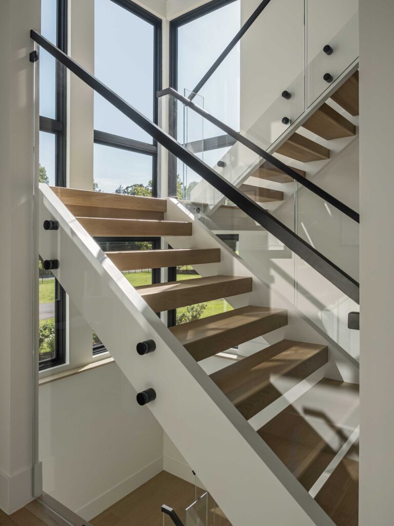 A side view of an open riser staircase with glass walls, black handrails and stained steps. Dark framed windows line the entire staircase.