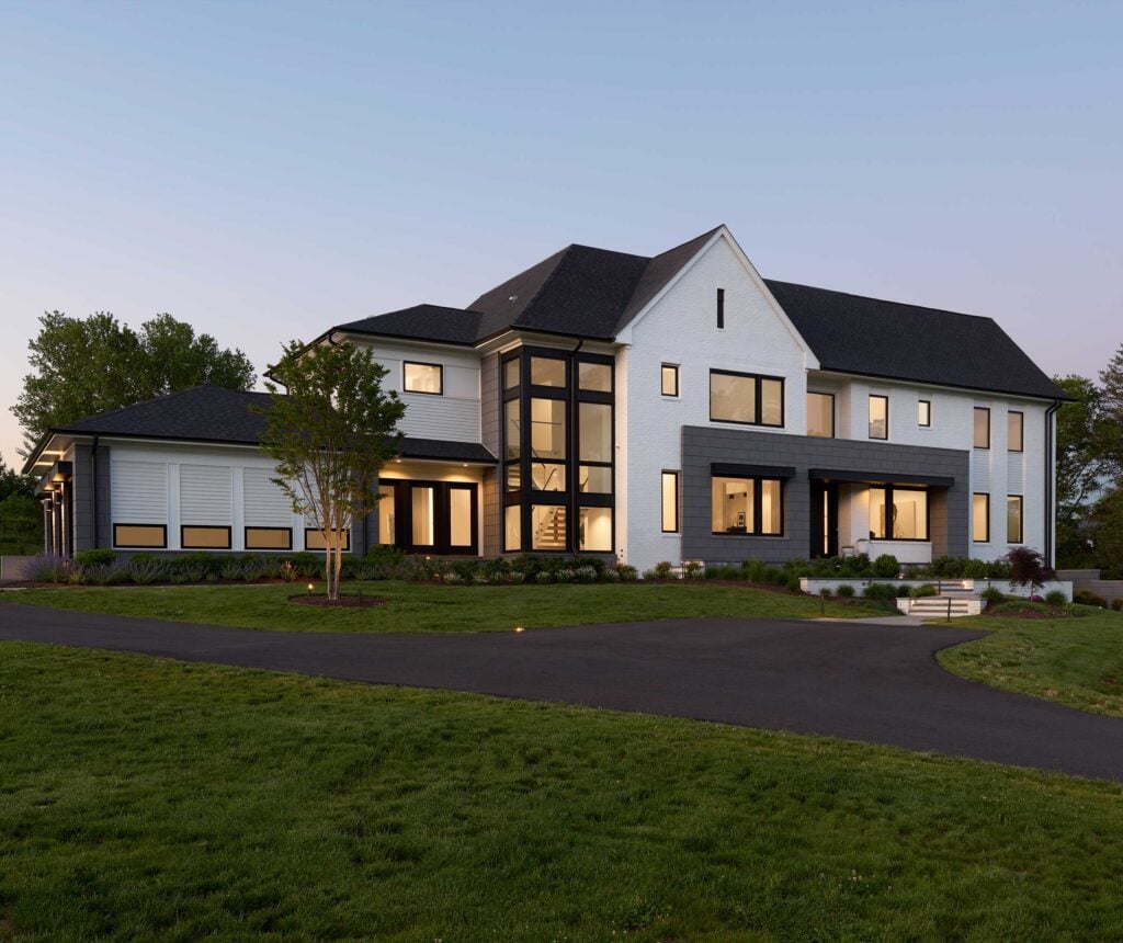 Front exterior image of a contemporary industrial style home in the early evening, sun setting, windows lit. house features geomteric shape of grey stone block, white brick and large windows including an exposed staircase.