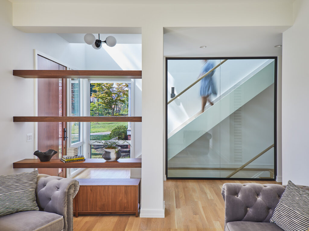 A set of three floating shelves separating the living room from the foyer, where the front door is open to the outside. To the right a blurred figure walks down a staircase behind a glass wall.