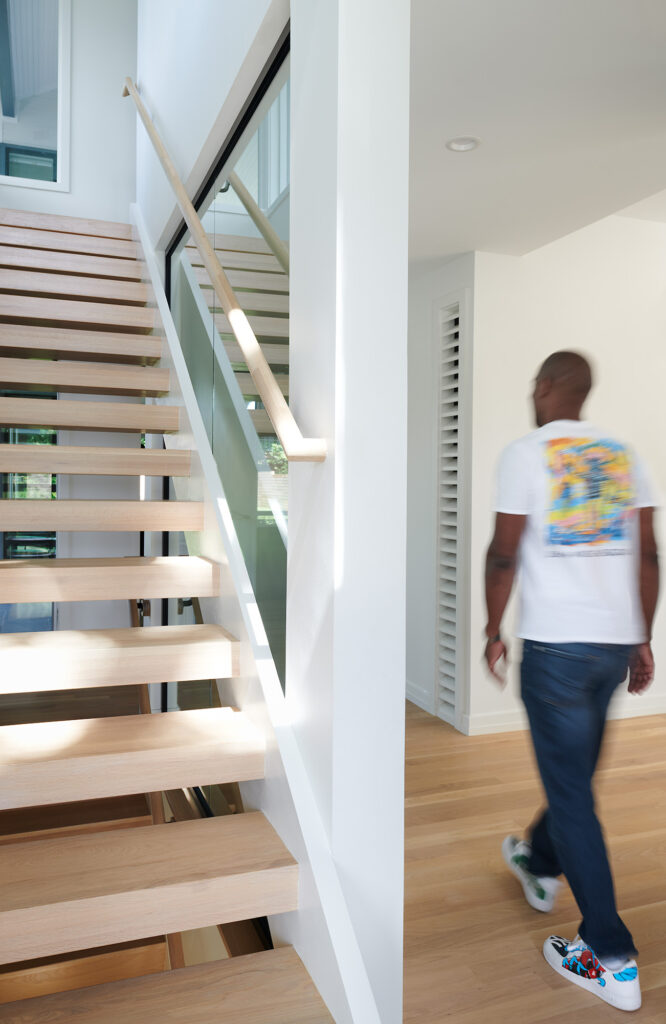 A man walks by an open riser staircase of light wooden treads with a wooden handrail along a large glass wall insert.