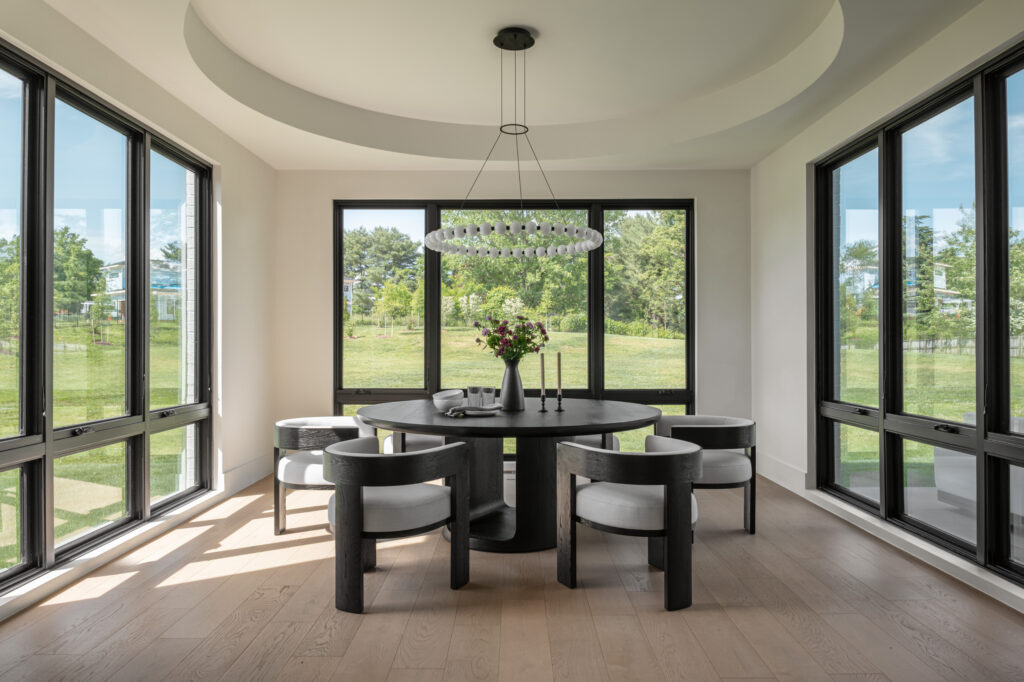 A morning room with a deep round tray ceiling, and round chandelier over a round table. The room is surrounded by nearly floor to ceiling windows on three sides. A sunny day and green grass outside.