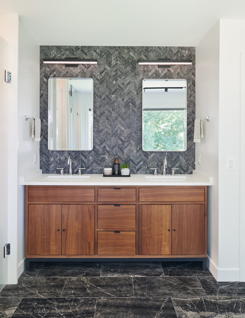 A double stained wood vanity with accent wall of Herringbone tile pattern in greys with specs of white, two mirrors, each with a light above.