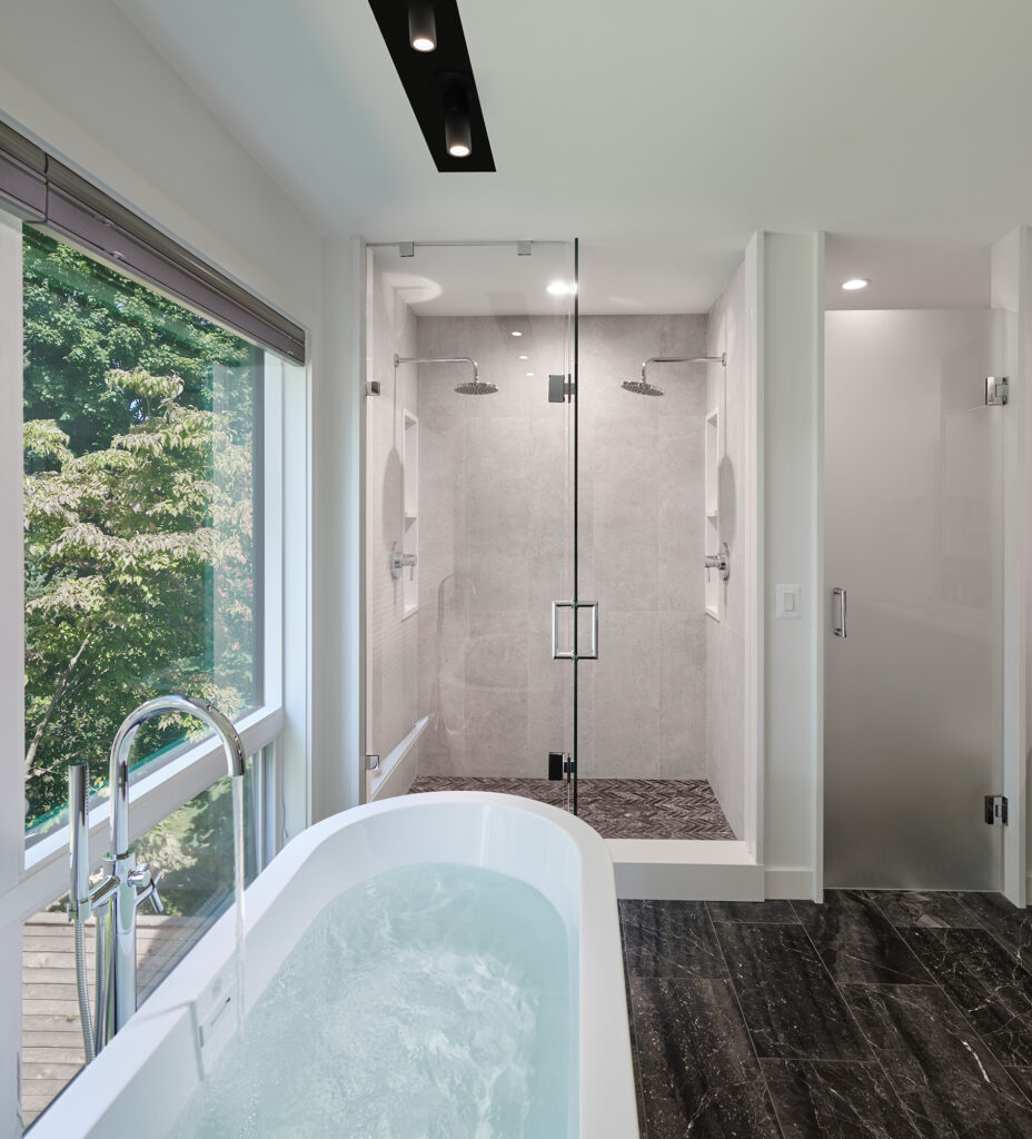A standing tub fills with water against a large grill-less picture window, a frameless shower behind it with dual shower heads, black tile floors.