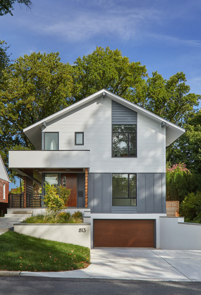 A head on view of a contemporary home with white siding and grey panels, unique windows and entry features in stained wood, exposed staircase and modern wraparound portico and drive under garage.