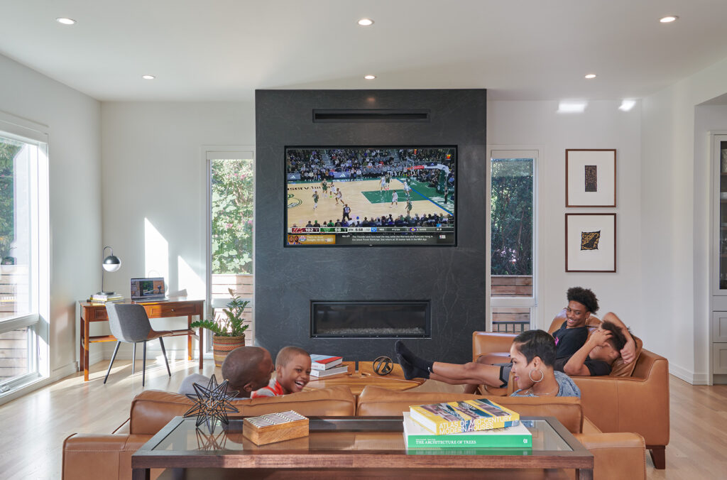 A family in their contemporary Family Room, a basketball game is on a television above a linear fireplace on a dark tile surround, leather couches tall modern grilless windows.