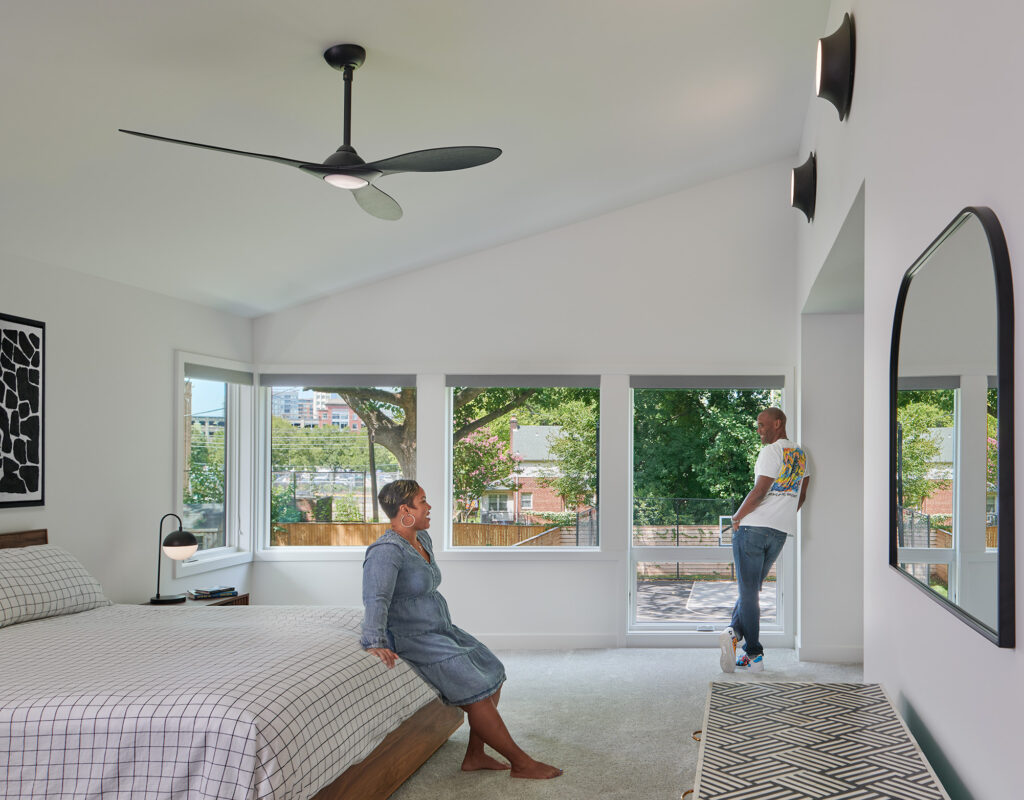 A large bedroom with oversized grilless windows open to the backyard, a man looks back from the windows at a woman sitting on the edge of the bed.