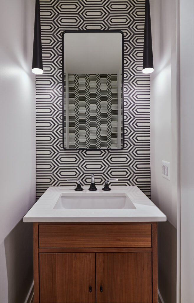 A contemporary powder room with patterned wallpaper reflected in the mirror, long black cone shaped lights hang to either side.