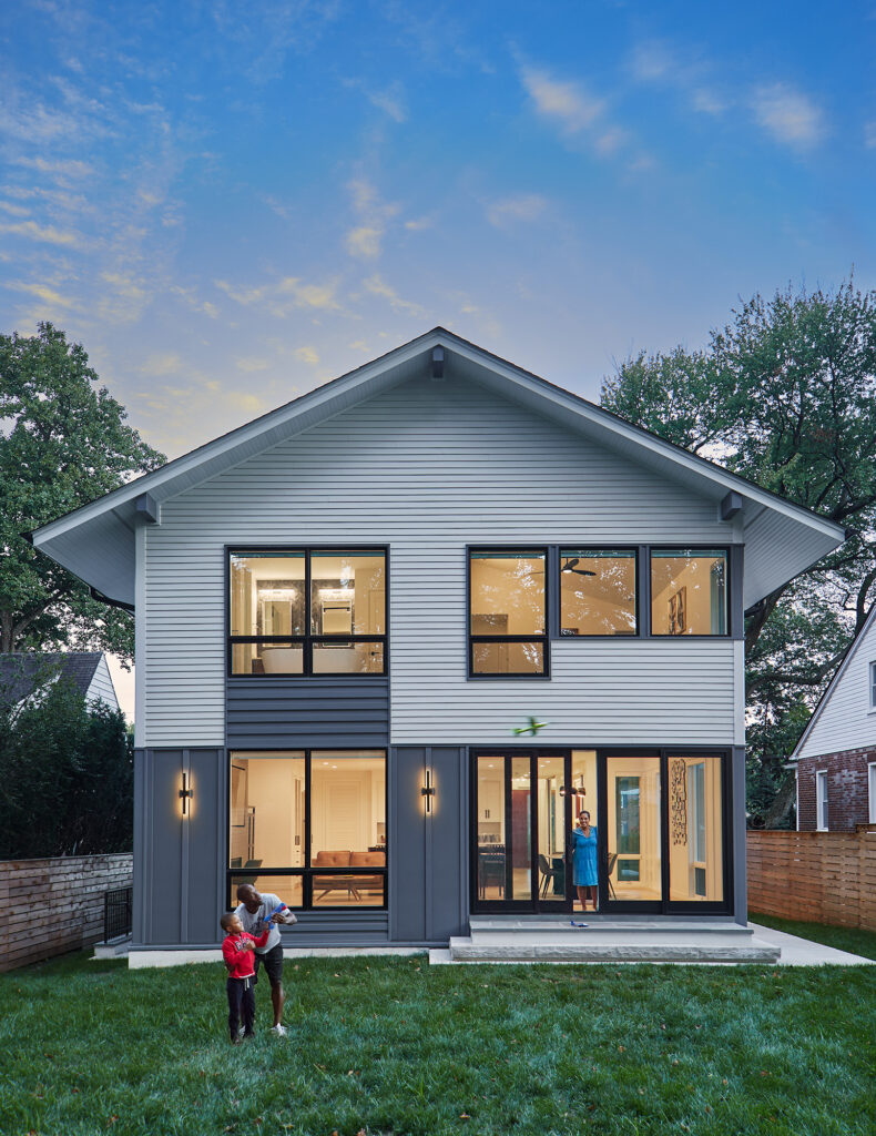 Looking at the back of a contemporary home from the yard, a man looks at the sky with a child, white siding with grey accent panels, lights on through modern grilless windows.