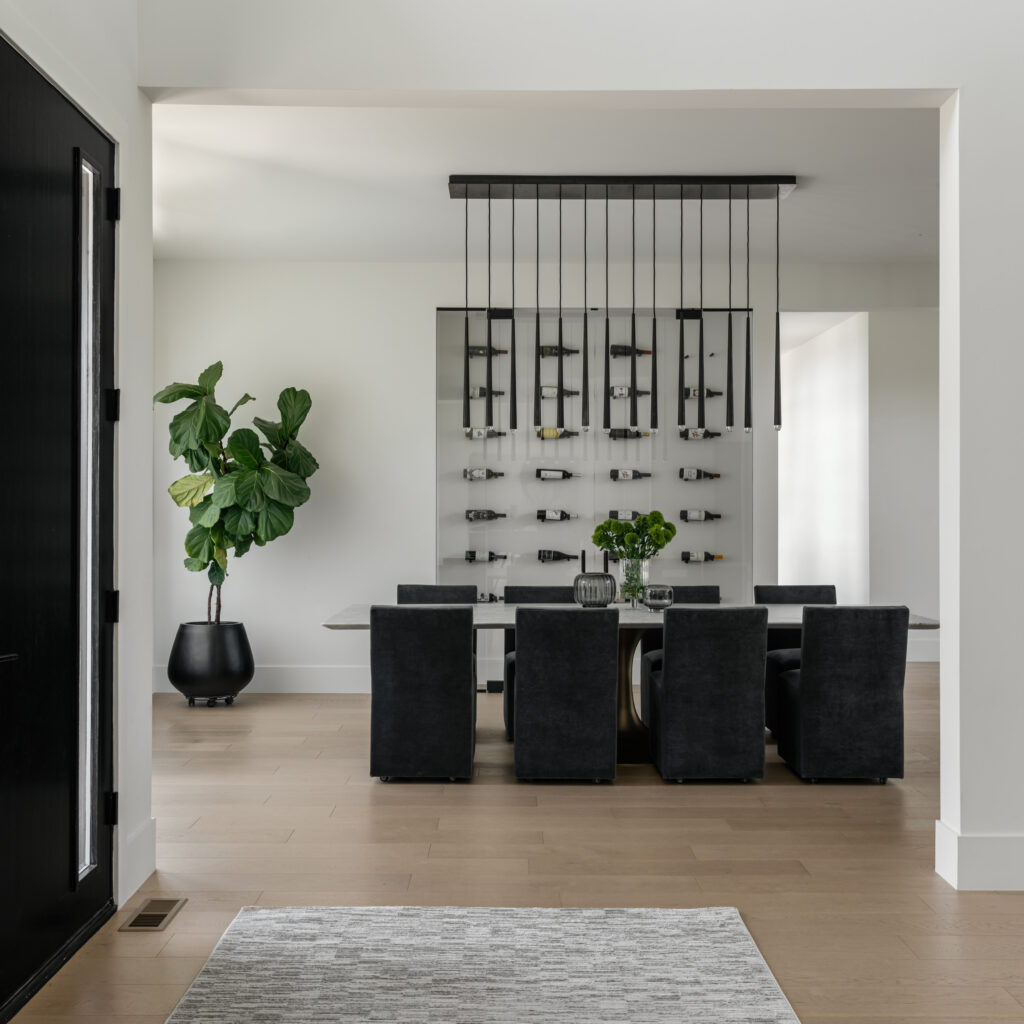 Looking into a dining room from the foyer, a dark chandelier and dark chairs contrast against a white room with light wood floors and wall display of horizontal wine bottles behind glass doors.