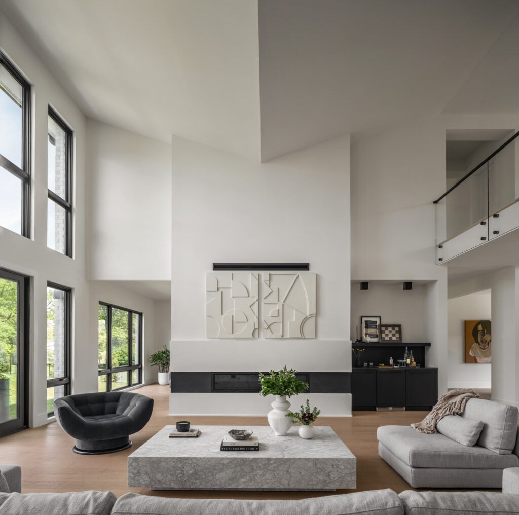 A two story family room with a vaulted butterfly ceiling, second floor overlook, linear fireplace and dry bar. Contemporary art hangs over the fireplace, tall windows to the left.