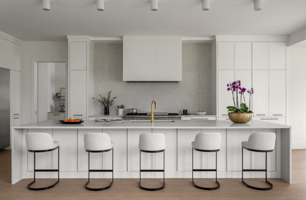 A naturally lit wide kitchen shot, white contemporary cabinetry, quartz waterfall island and backsplash, white fume hood cover. Stainless steel appliances and black iron base stools and purple orchids add color.