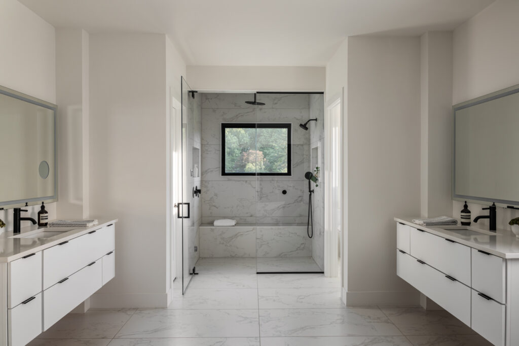 An owner's suite bath with dual floating vanities on either side in the foreground, white cabinetry and counterops, black fixtures. Beyond a glass walled shower with wall and overhead showerheads and quartz wall and seat.