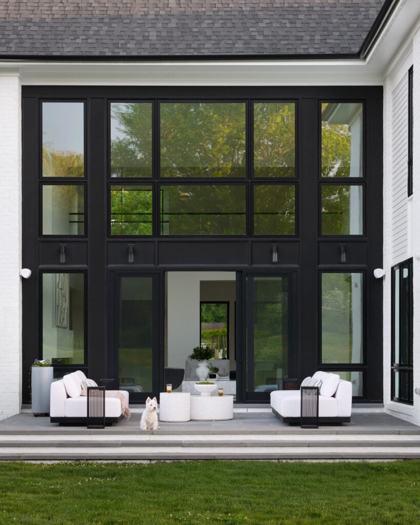 A rear patio photo, white couches and tables, dark side tables and dark frame oversized windows with sliding doors open to inside.