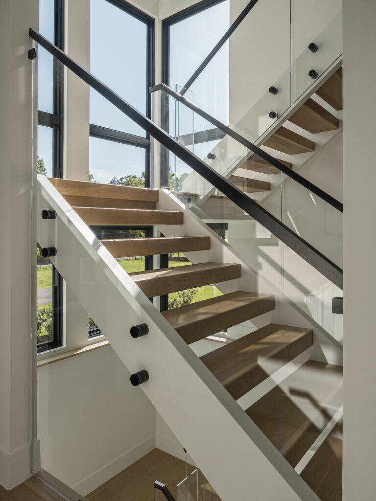 A side view of an open riser staircase with glass walls, black handrails and stained steps. Dark framed windows line the entire staircase.