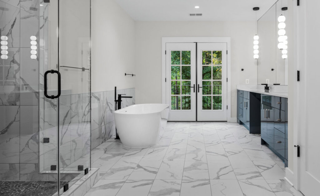 An owners bath with frameless glass enclosed shower in the left foreground, a free standing tub with black tub filler beyond it, a double vanity to the right and doors at the end of the room to an outdoor romeo and juliet balcony.
