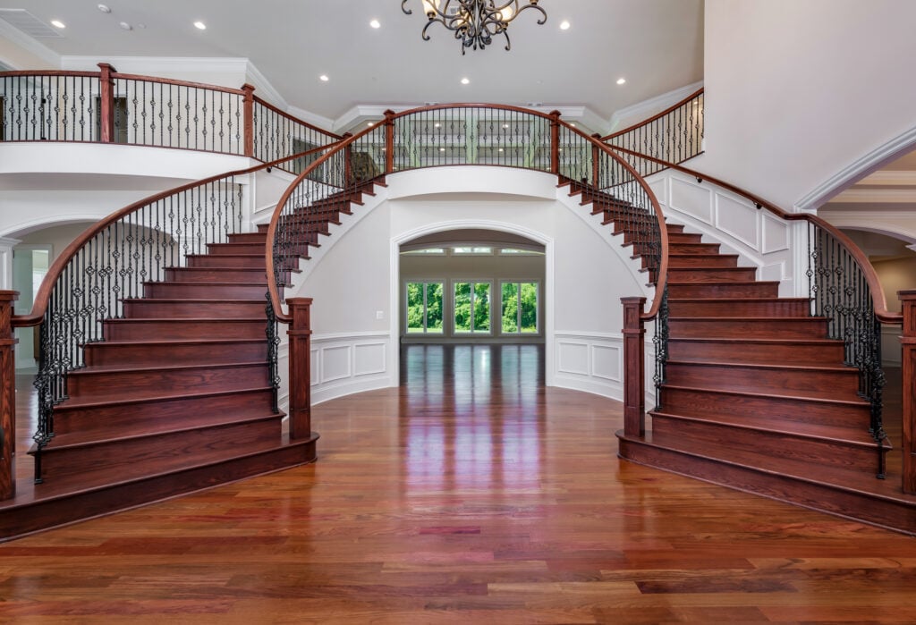 A dual curved staircase in a dark reddish stained wood, with iron black vertical balusters, wooden newel posts and wooden rails. Beyond a two story family with a coffered ceiling is visible.