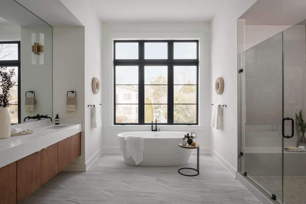 An Owner's Bath with free standing tub in front of large dark picture window, frameless shower to the right, double vanity to the left
