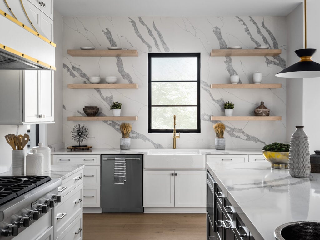 An accent wall of Q Premium Natural Quartz with floating natural wood shelves, farmhouse-style sink, and matching countertop. Quartz kitchen island and professional cooktop in the foreground.