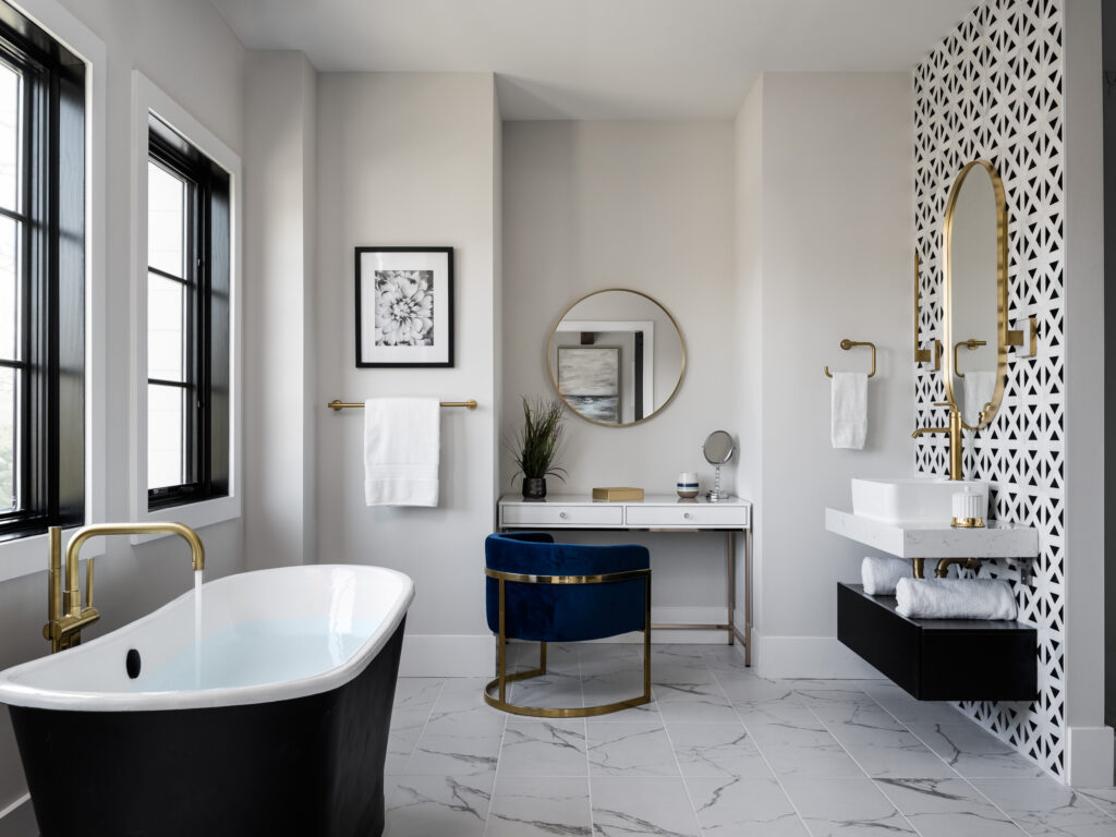 An Owners bath with freestanding tub in foreground, filling with water, black outside, white inside. Vanity and make up desk visible.
