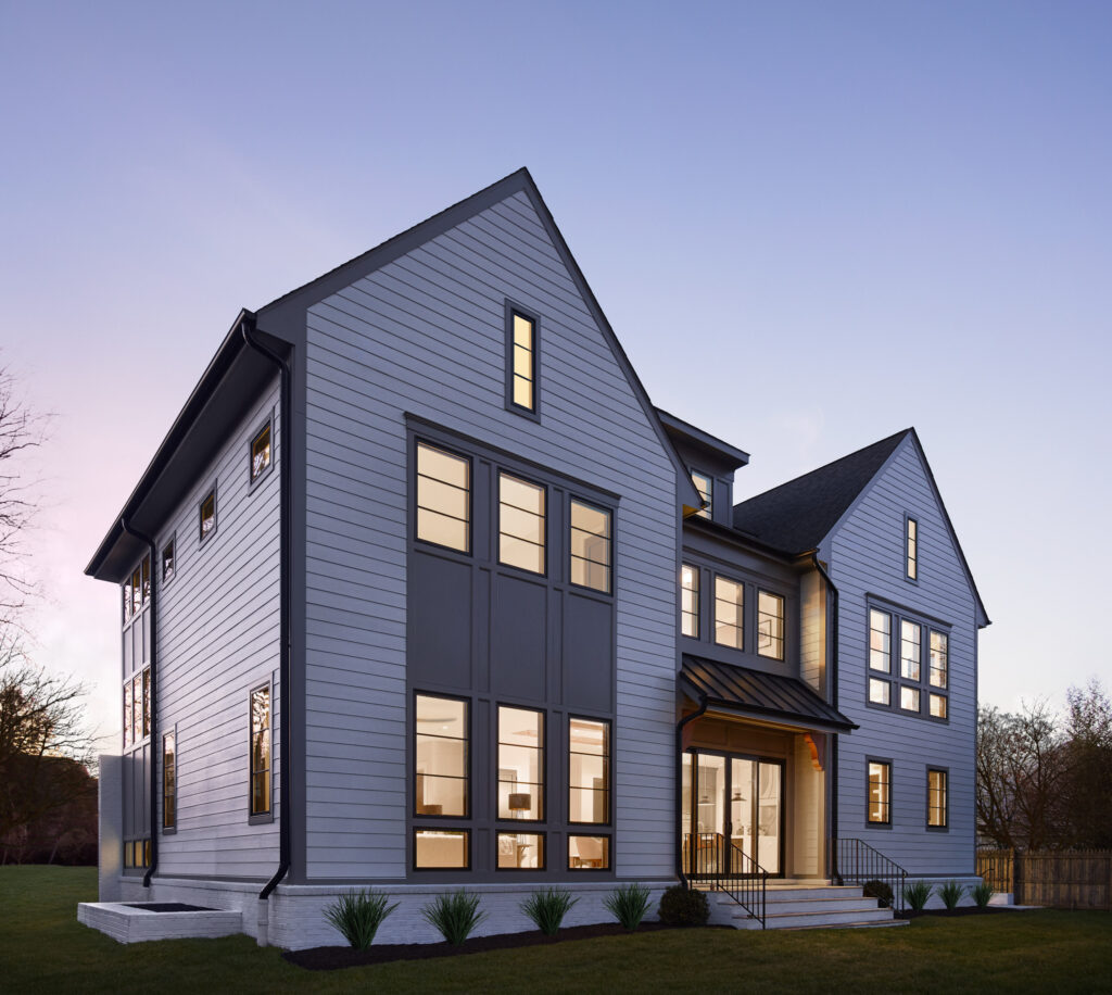 An angled twilight shot of the rear of a home lit up against the darkening sky. Home features white panels, grey accents and porch with overhang and stairs to grade.