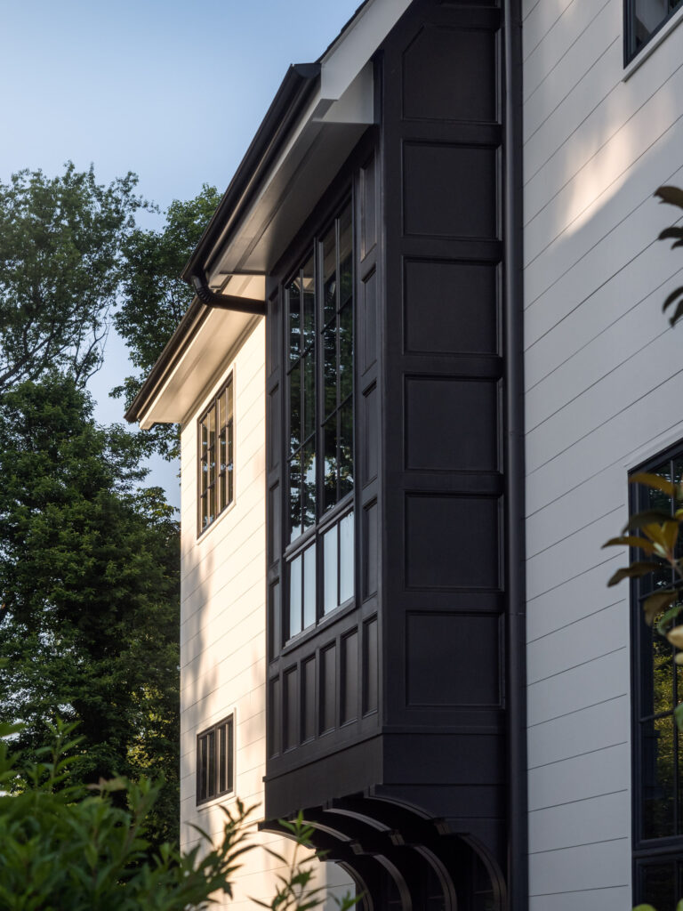 Close up of a tall bay window assembly of of dark panels extrudes from a white plank wall.