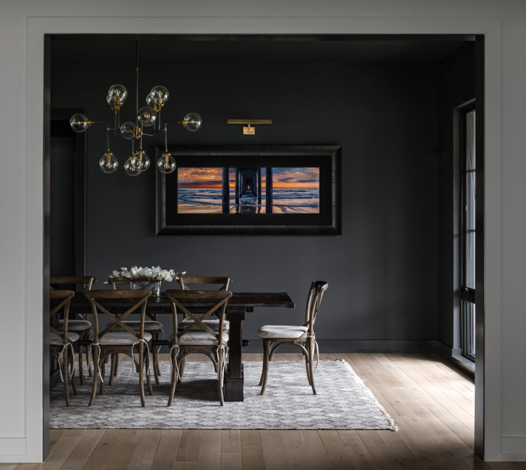 A dining room with dark accent wall and colorful artwork, table and chairs in the center, modern chandelier of glass globes, natural light pours in from the right.