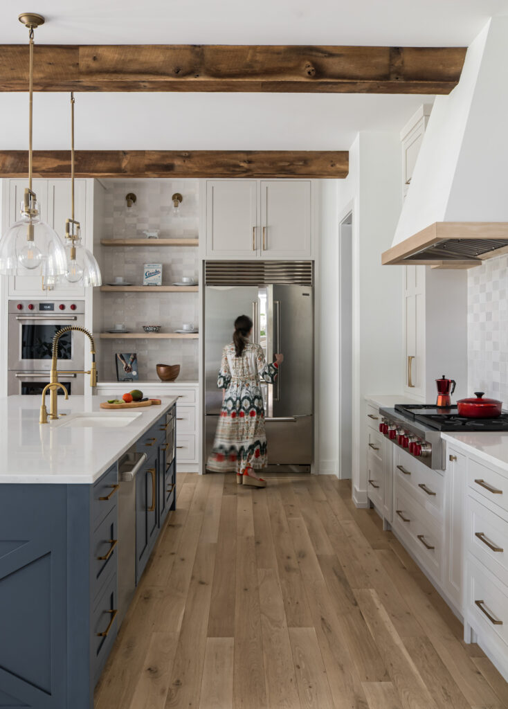 A woman stands at the refrigerator from the far side of a modern farmhouse kitchen with blue island, exposed beams, tile backsplash and accent wall