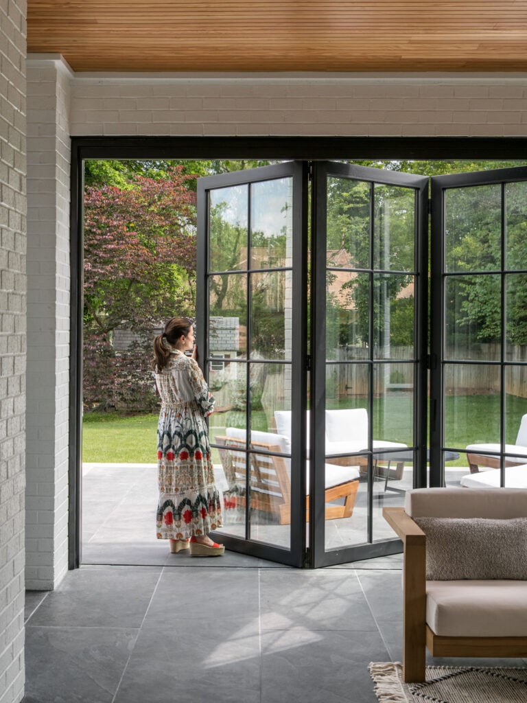 A woman stands at a folding patio door, opening to a patio and green backyard.