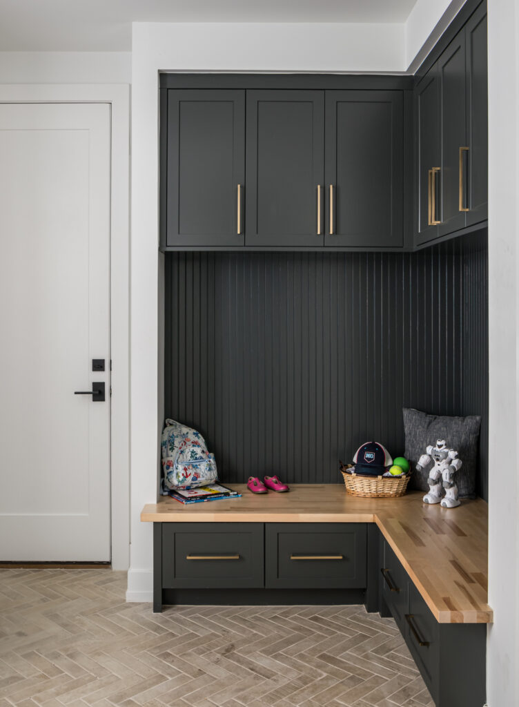A mudroom with natural wood bench, dark paneled wall and dark cabinets, children's toys sit on the bench seat.