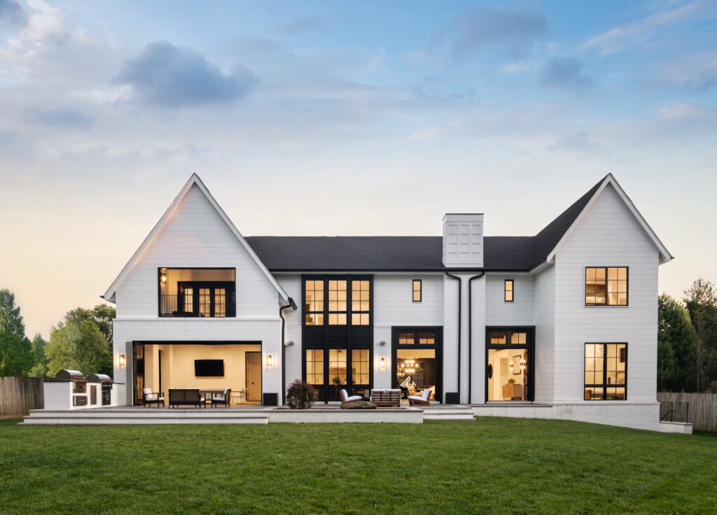 The rear of a custom modern farmhouse with an indoor outdoor space open the terraced rear patio, additional exterior doors also open, with lights in all windows against a darkening sky.