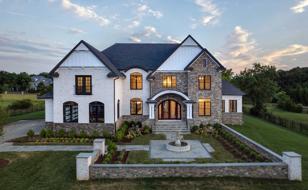 A home in white powdered brick and natural stone, with black windows and highlights, white board and batten in the gables and a wide curved portico.