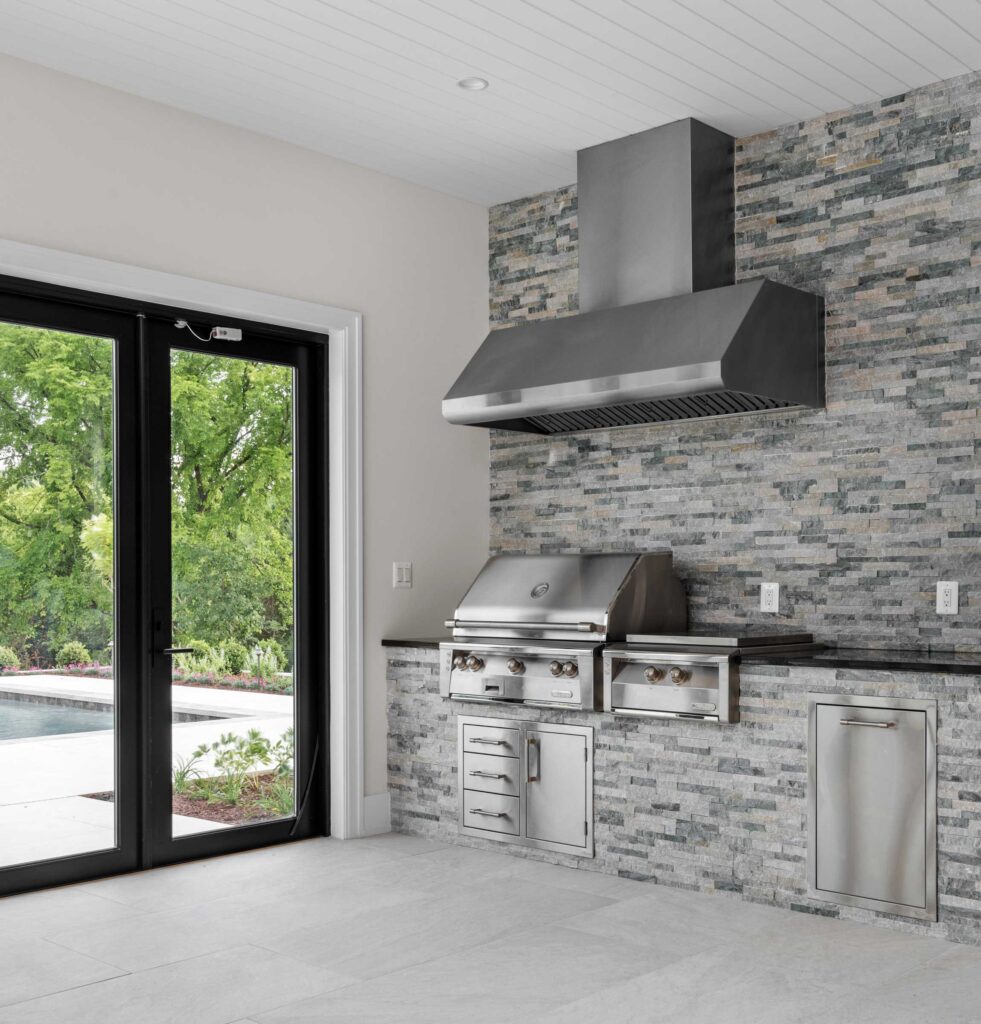 A room with a wall of glass doors to a pool and pool deck visible outside. Along a stone accent wall is a grill under a large fumehood with a cooktop, counter, sink, mini fridge and various storage compartments.