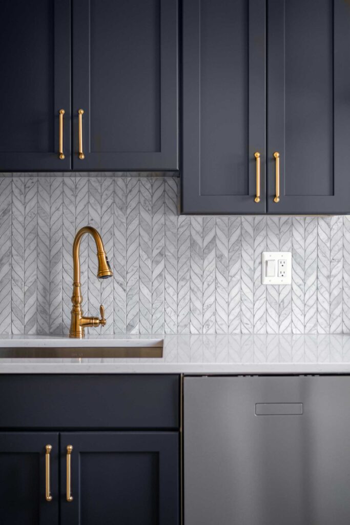 A close up of blue cabinets with brass handles, a white coutnertop and brass faucet with intricate tile backsplash.