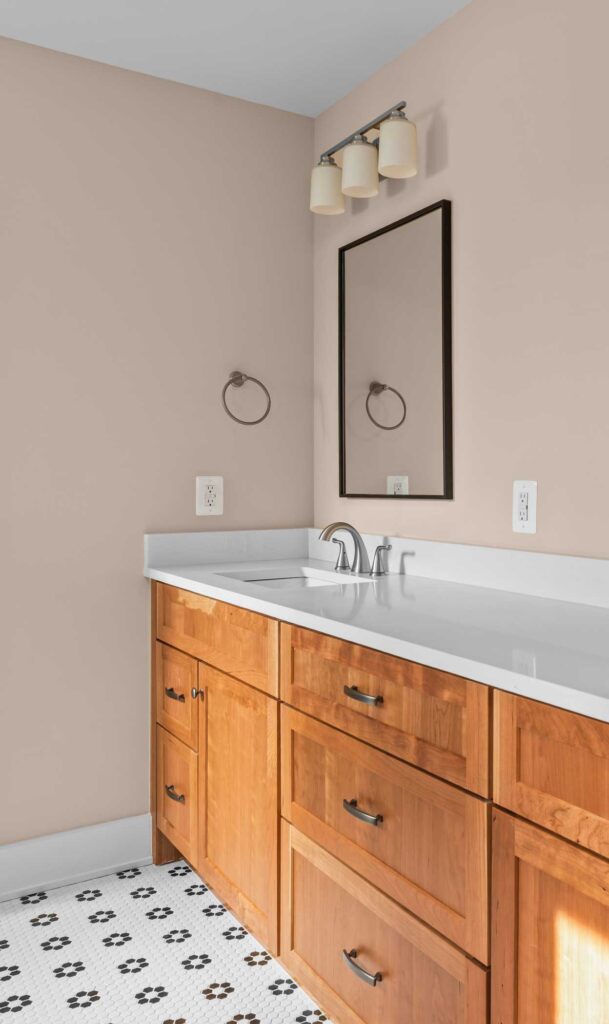 the corner of an owner's suite bath with long white quartz counter and stained wood cabinet base with a hand crated look and dark hardware. Dark framed mirror and light above.
