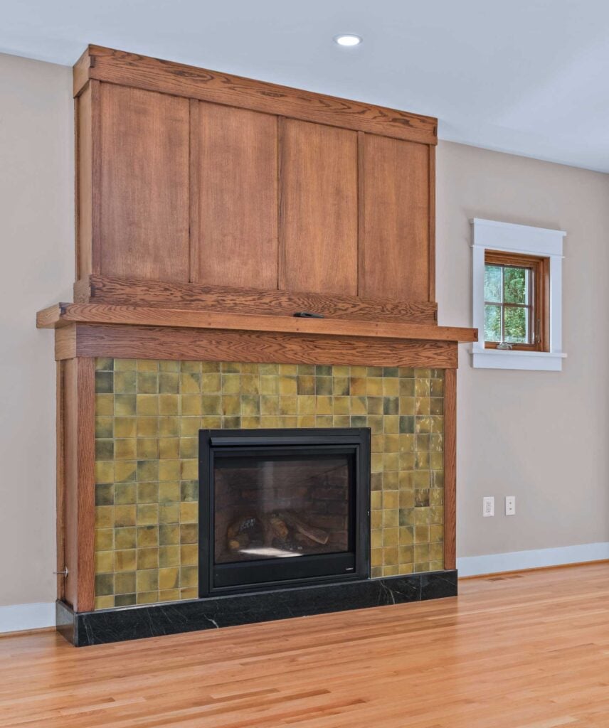 A square gas fireplace surrounded by square tile in greens and browns, framed with stained wood. Stained wood panels above.