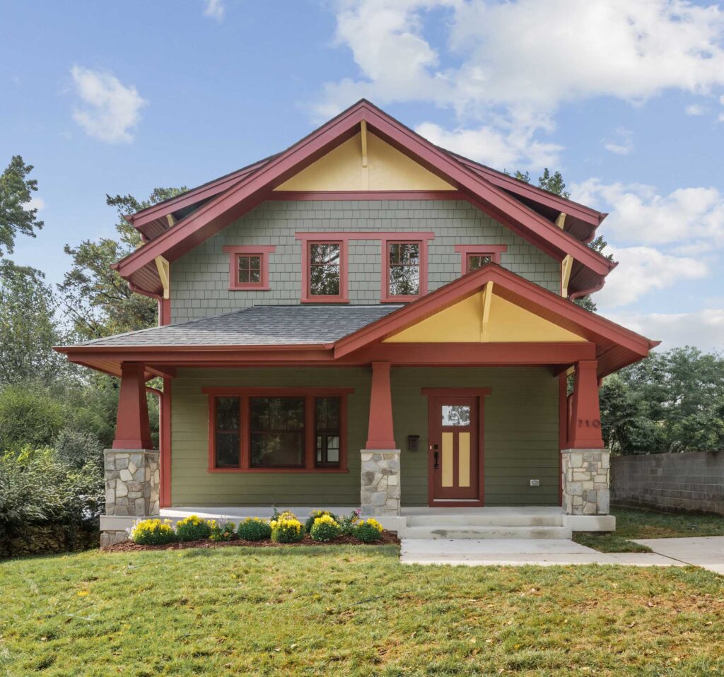 A craftsman style front home exterior in green hardie shingles with red columns, fascia and window trim, and yellow accents and brackets. Green grass, blue sky.