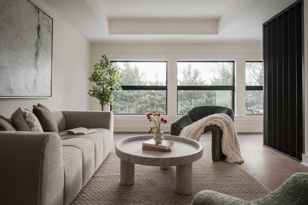 A loft with deep tray ceiling and black slatted accent wall divider, a couch to the right, round table in the center, chair behind table, row of modern dark windows with trees outside.