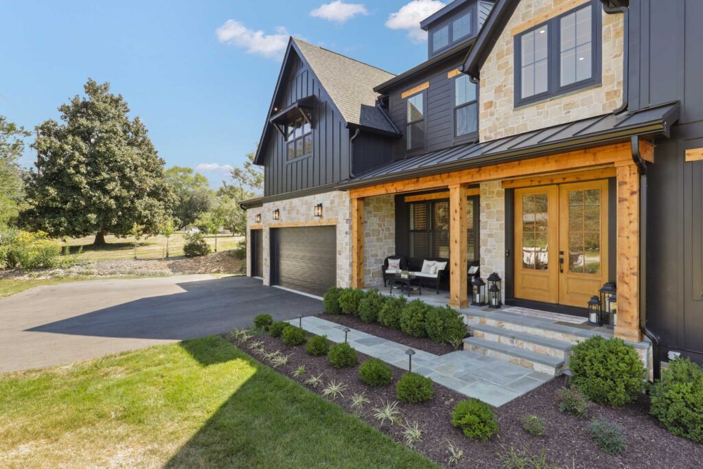 A angled view of the front of a farmhouse style home with the front door in the foreground. Espresso colored board and batten mixed with stone and stained wood posts, door and accents. A sunny day, green lawn, trees and bushes.