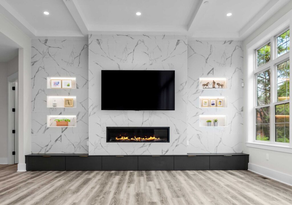 A family room with a quartz accent wall with gas linear fireplace beneath a flat TV, two columns of three niches cut into either side of the wall, coffered ceiling, hardwood floor, windows to the right.