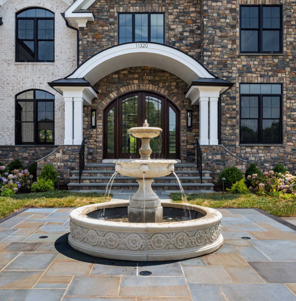 A circular fountain in front a home, sitting on a multi color flagstone front patio area, stone riser stairs to the front door under a wide curved portico with double posts. The home is stone and white brick.