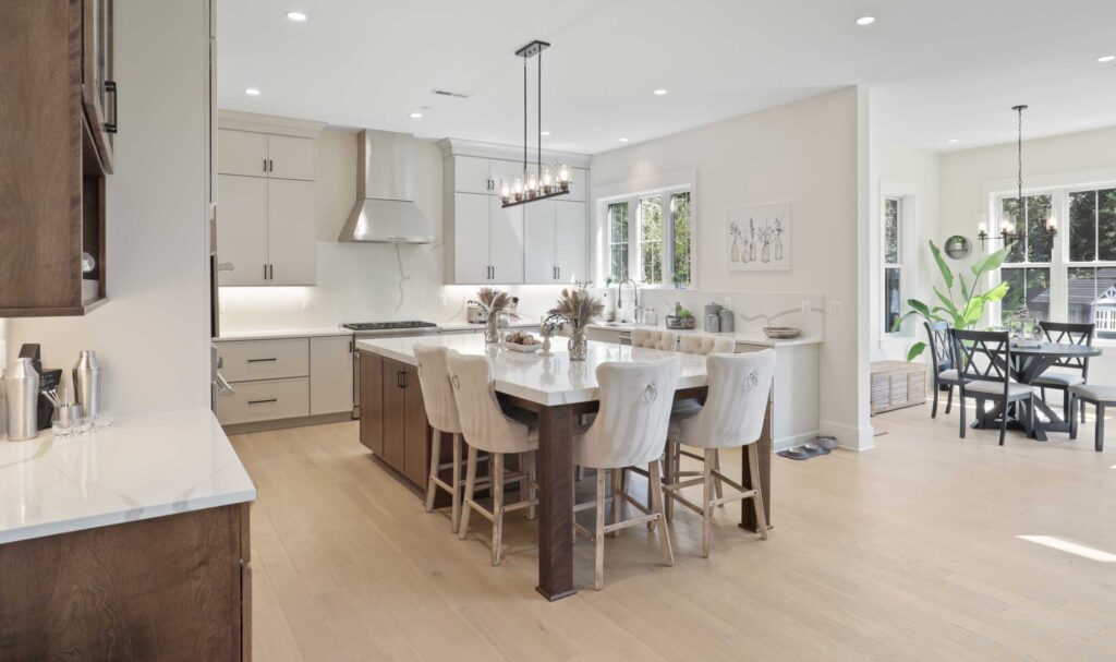 A kitchen with an island front a center, 6 chairs seated around a table like section, followed by cabinets. White quartz top, stained brown wood base and legs. Behidn the kitchen has white cabinets and stainless steel appliances.