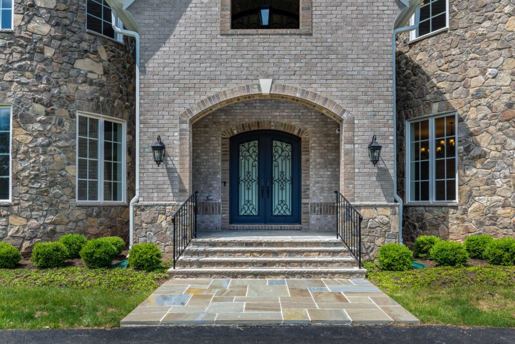 A brick portico with curved entryway and curved iron double door, carriage lights on either side, flagstone stairs with stone risers to the landing, stone on either side of the portico