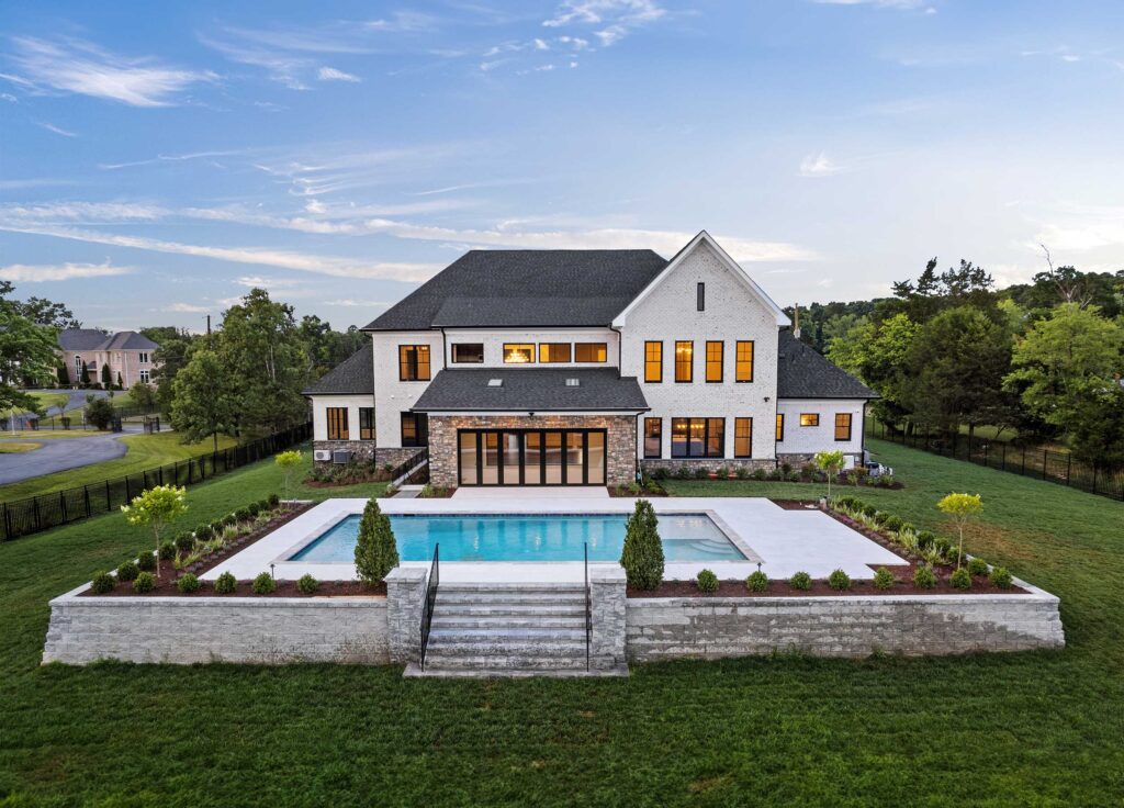 A photo of the rear of a home in white powdered brick with a stone lounge area with wide glass doors opening to a pool. The pool is raised, surrounded by stone wall and stairs up to the pool deck.