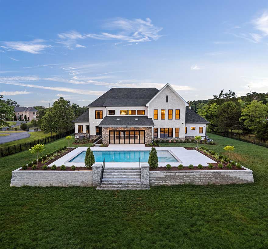 A drone photo of a the rear of a home lit up against an early evening sky, in the foreground a pool surrounded by a stone wall stepped up from the yard