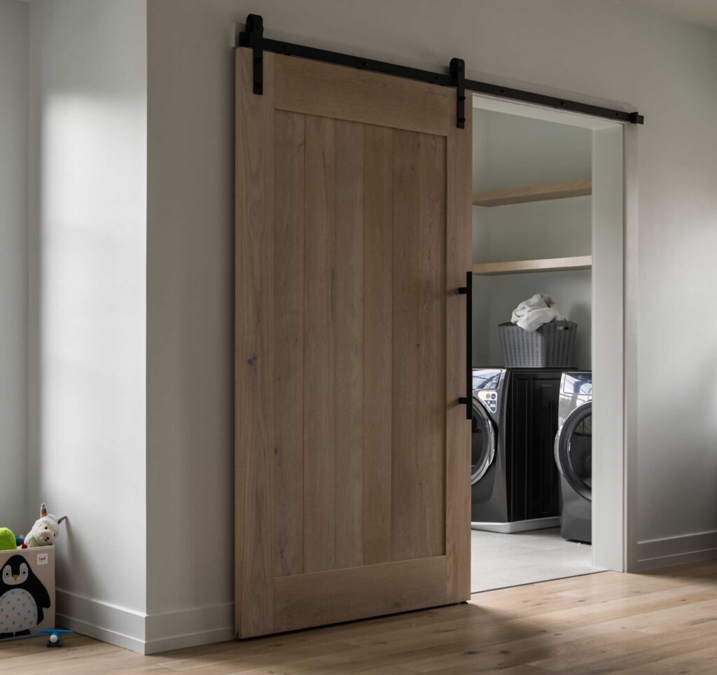 A washer and dryer behind a sliding stained wood barn door with black hardware, matching wood shelves above the machines, children's toys in the left foreground
