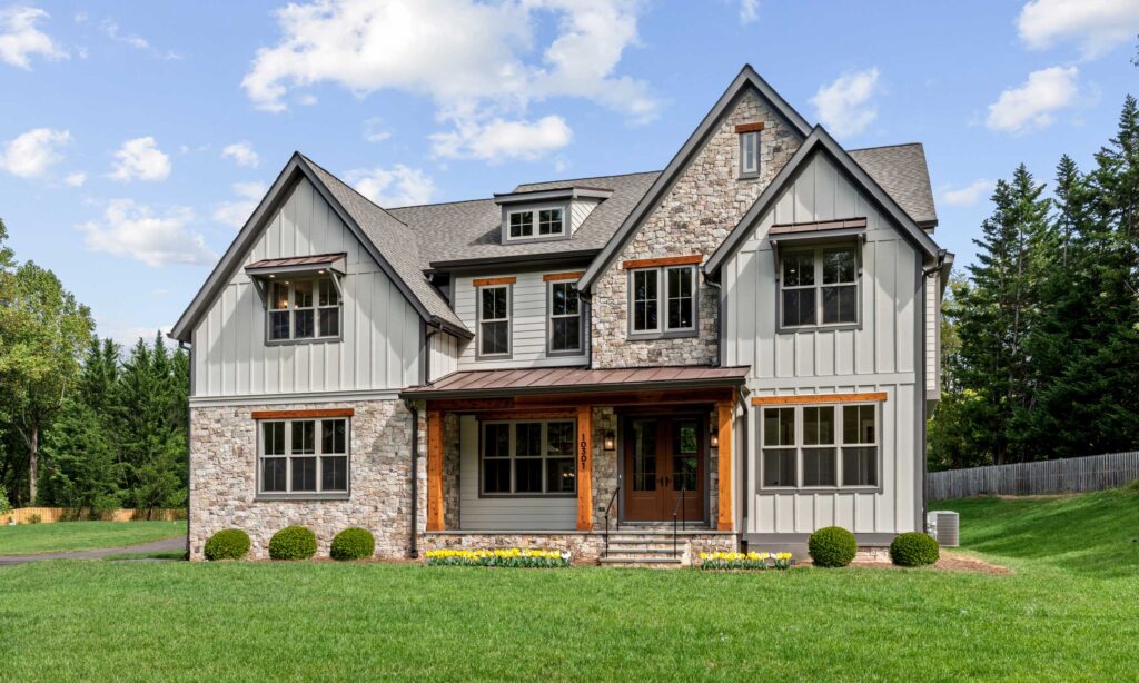 A modern farmhouse style front exterior in beige board and batten, with matching stone and stained posts and accents. A double revers gable on the left. Home sits in a grassy field with forest in the background.