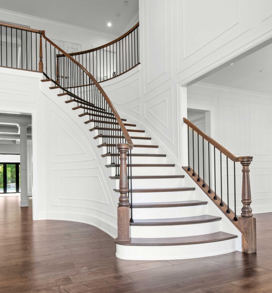 A curved staircase with stained wood treads, white painted risers, wood posts and rails, black iron ballusters, and shadow box details on the walls.