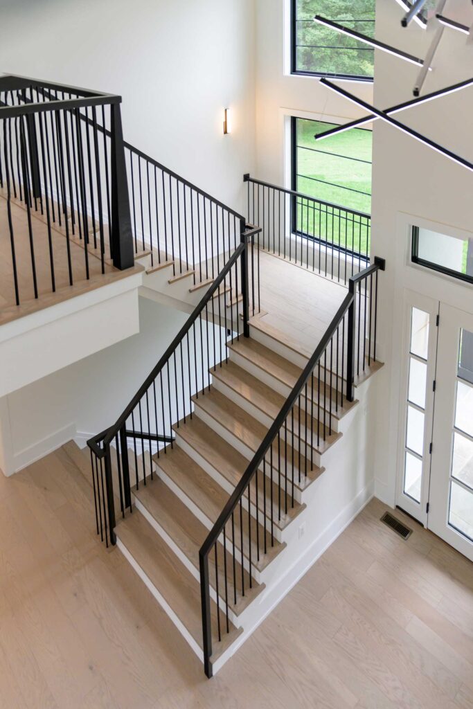 Looking down from a landing above at a staircase with stained treads and white painted risers, black vertical rails. A very contemporary chandelier hangs from the ceiling of this two story space. Large windows provide lots of natural light.
