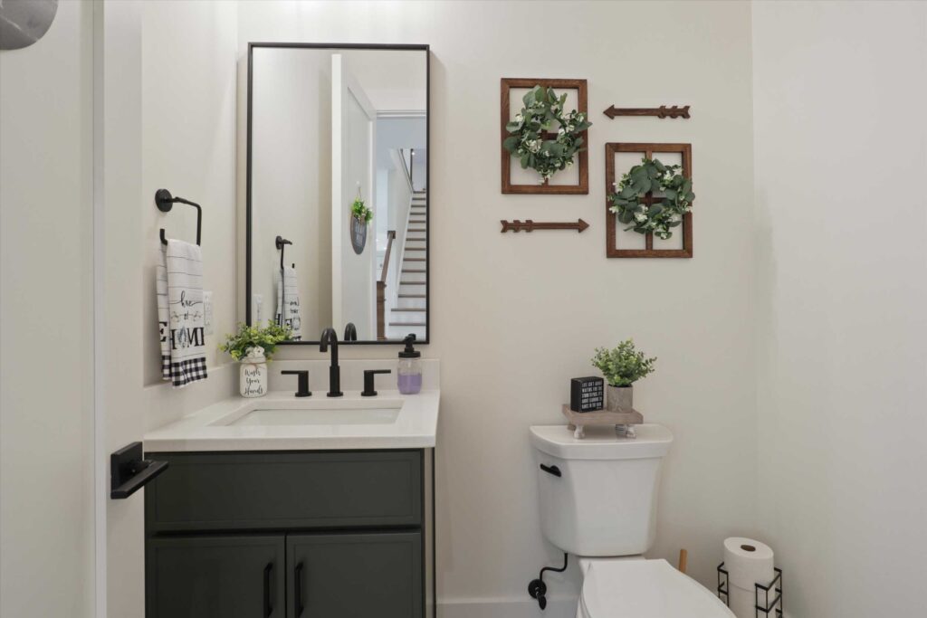 Powder room featuring a dark grey vanity with a white countertop, a black-framed mirror, and farmhouse-style wall decor above the toilet.