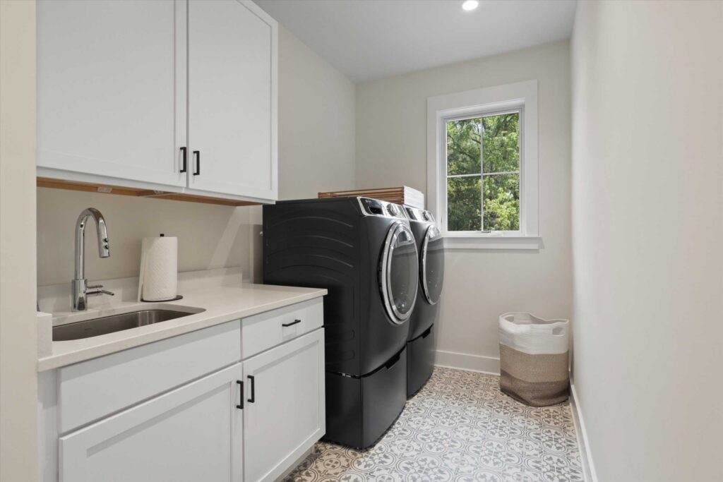 Laundry room equipped with black front-load washer and dryer, white shaker cabinetry, a stainless steel utility sink, and patterned floor tiles.
